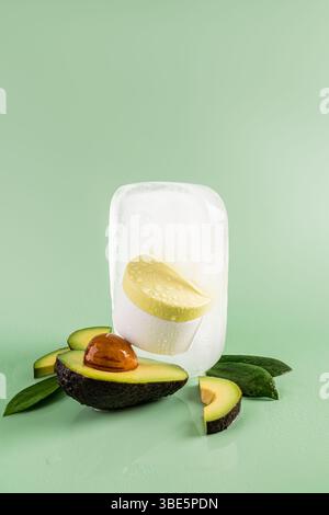 Frische Avocadoblätter und Hautpflegeprodukte in einem klaren Eisblock, umgeben von geschnittenen Avocados auf einem glatten grünen Hintergrund, die eine lebendige, raffinierte Atmosphäre schaffen Stockfoto