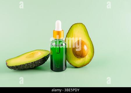 Eine grüne Flasche Avocadoöl mit einem Tropfer, zwischen zwei frischen Avocadohälften auf hellgrünem Hintergrund platziert. Perfekt für Schönheit, Wellness und h Stockfoto