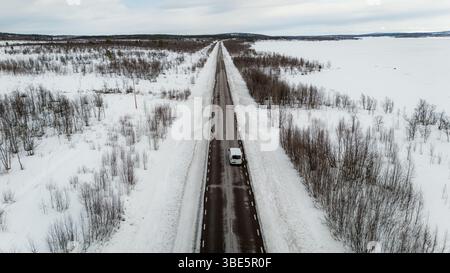 Aus der Vogelperspektive auf einer langen, geraden Straße, die durch eine schneebedeckte Landschaft führt, gesäumt von kargen Bäumen unter einem bewölkten Winterhimmel, die den Essens einfangen Stockfoto