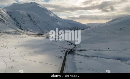 Aus der Vogelperspektive zeigt eine atemberaubende nordische Landschaft mit einer kurvenreichen Straße, die durch weite, schneebedeckte Berge unter einem bewölkten Himmel führt. Ideal für Stockfoto