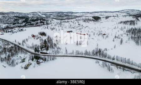 Klare Aussicht aus der Luft auf eine schneebedeckte nordische Landschaft mit schneebedeckten Feldern, verstreuten Bäumen und roten Häusern unter einem weitläufigen bewölkten Himmel. Straßenkurve Stockfoto