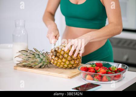 Eine schwangere Frau in einem grünen Yoga-Outfit schneidet eine frische Ananas auf einem Holzbrett. Erdbeeren in einer Schüssel und ein Krug Milch sind auf dem weißen Kitc Stockfoto