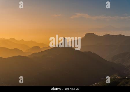Ein bezaubernder Blick auf den Sonnenuntergang von Roque Nublo, der sich vor dem Hintergrund mehrschichtiger Bergsilhouetten in warmen Gold- und Orangetönen abhebt Stockfoto