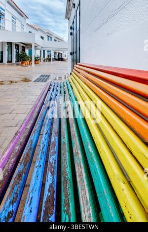 Lebendige, regenbogenfarbene Holzbank, die sich in Eine moderne urbane Umgebung ausdehnt. Farbenfrohe Öffentliche Sitzplätze, Perspektivische Sicht, Zeitgenössische Architektur Stockfoto