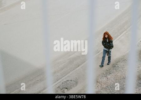 Person mit rotem Haar, die in lässiger Kleidung entlang der Stadtstraße geht, teilweise durch Bars aus der Sicht von oben gesehen, was eine düstere Atmosphäre verleiht Stockfoto