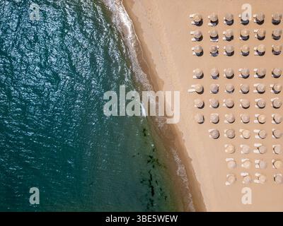 Von oben aus blickt man auf einen Sandstrand mit Strohschirmen und Liegen, die in Reihen angeordnet sind, und Wellen, die sanft an Land waschen. Sommerferienkonzept Stockfoto