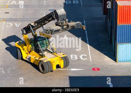 Gelber REACH-Stapler, der sich zwischen Versandcontainern im Frachtterminal bewegt Stockfoto