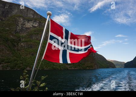Eine majestätische norwegische Flagge weht vor dem Hintergrund atemberaubender Fjorde und eines klaren blauen Himmels während einer Bootsfahrt. Stockfoto