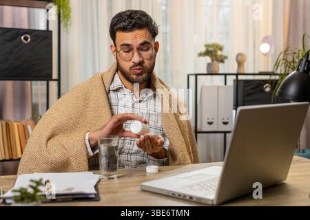 Gesundheit, Kälte und Menschen Konzepte. Der kranke, junge indische Geschäftsmann, in Decke gehüllt, arbeitet auf dem Laptop am Schreibtisch im Heimbüro. Ungesunder Hispanic Freelancer nimmt Medikamente und zittert vor der Kälte zu Hause. Stockfoto