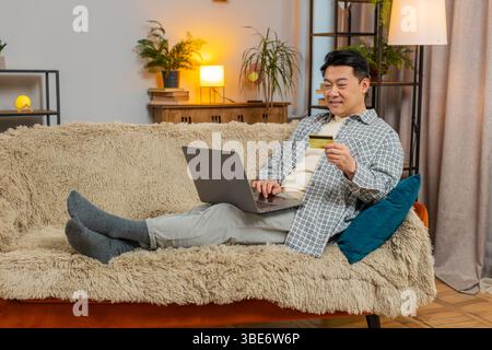 Glücklicher Mann mittleren Alters mit Kreditkarte und Laptop beim Überweisen von Geld, kauft Online-Shopping bargeldlose Bestellung Lebensmittel Lieferung zu Hause Wohnung drinnen. Der Chinese sitzt auf der Couch. Stockfoto