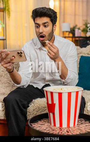 Indischer Mann auf dem Heimsofa isst Popcorn, während er ein Video auf dem Smartphone ansieht, voll mit Inhalten beschäftigt. Arabian Guy genießt Entspannung, eingetaucht in digitales Entertainment und erlebt Spaß und Spannung Stockfoto