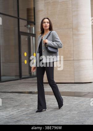 Eine Frau in einem grauen Blazer und einer schwarzen Hose geht selbstbewusst draußen. Sie trägt eine schwarze Handtasche und trägt schwarze Absätze. Glasfenster und ein großer Stein Stockfoto