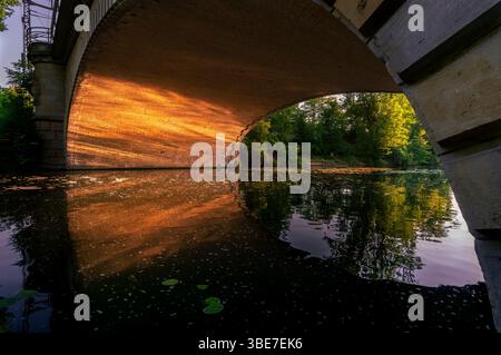 Steinbogenbrücke über den Landwehrkanal in Berlin, glühend im goldenen Stundenlicht mit Reflexionen auf dem ruhigen Wasser und üppigem Grün im Hinterland Stockfoto