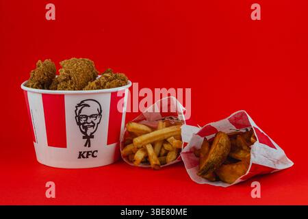 Lemberg Ukraine 05.21.25 Ein Eimer mit knusprigem gebratenem Huhn wird von zwei Portionen goldener Pommes frites und Kartoffelscheiben begleitet, die alle gegen einen bo gestellt werden Stockfoto