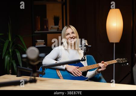 Lächelnde Musikerin singt und spielt Akustikgitarre, während sie einen Song in einem Heimstudio aufnimmt Stockfoto