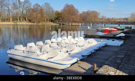 Trakai Litauen - 03.28.2025 Weiße Tretboote stehen auf einem hölzernen Dock neben einem ruhigen See mit roten Ruderbooten und einer hölzernen Brücke im Hintergrund u Stockfoto