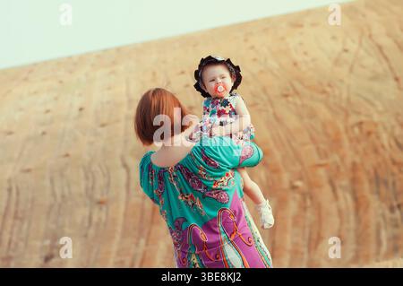 Mutter hält ihre kleine Tochter in Armen, steht draußen auf einem sandigen Hügel. Familienliebe, Sommerbindung und Elternschaftskonzept in natürlichem Licht Stockfoto
