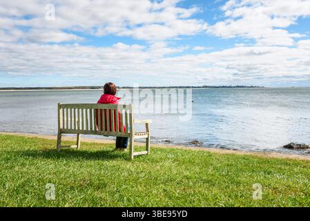 Frau, die auf einer Holzbank vor der Bucht sitzt, an einem schwachen Herbstmorgen Stockfoto