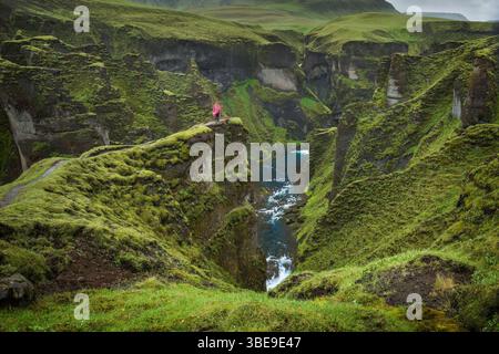 Blick auf den Fluss, der durch das Kanu Fjadrargljufur fließt, bedeckt mit grünem Gras und Moos auf Island dramatische Landschaft und eine Person in Regenmantel Stockfoto