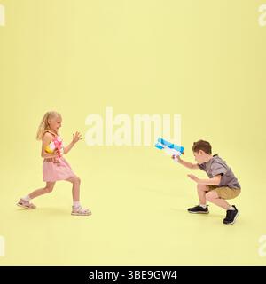 Mädchen und Junge spielen Wassergewehrkampf, Mädchen lächelnd, während sie rosafarbenes Spielzeug halten, energiegeladener Spaß in Bewegung vor pastellfarbenem Hintergrund Stockfoto