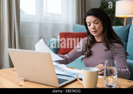 Fokussierte Frau, die von einem gemütlichen Wohnzimmer aus arbeitet und Datendiagramme auf Papier mit einem Laptop analysiert. Freie Geschäftsfrau Wirtschaftstätigkeit Stockfoto