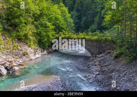 Malerischer Blick auf eine alte Steinbrücke über einen klaren türkisfarbenen Fluss, der durch einen üppig grünen Wald in Bregenzerwald, Vorarlberg, Österreich fließt Stockfoto