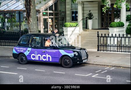 London, England, 12. Juli 2023, Ein Classic Black London Taxi mit leuchtender purpurpurfarbener Werbung parkt am Hoteleingang, während ein Passagier abgesetzt wird Stockfoto