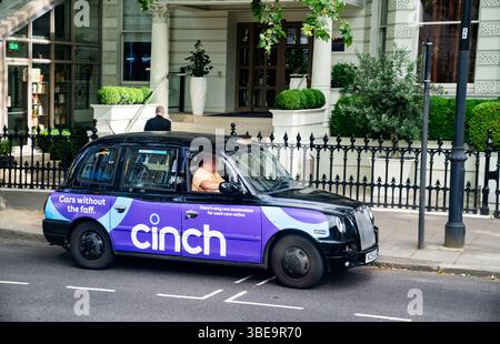 London, England, 12. Juli 2023, Ein Classic Black London Taxi mit leuchtender purpurpurfarbener Werbung parkt am Hoteleingang, während ein Passagier abgesetzt wird Stockfoto
