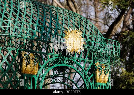 Dekorativer grüner, schmiedeeiserner Pavillon mit goldenen Sonnen- und Laubmotiven, vor bewaldetem Hintergrund im frühen Frühjahr oder Spätwinter. Stockfoto