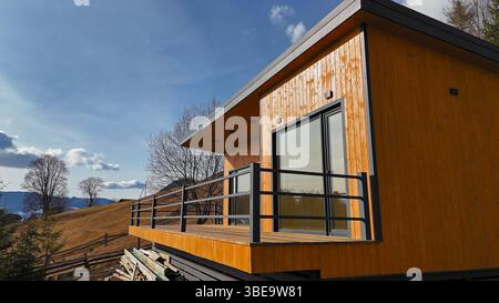 Moderne Holzhütte mit malerischem Bergblick. Eine moderne Holzhütte mit großen Glasfenstern und einem Balkon in einem malerischen Hügel Stockfoto
