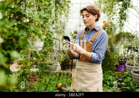 Eine junge Frau pflegt ihre Pflanzen in einem Gewächshaus und verwaltet ihre Gartenfläche nachdenklich. Stockfoto