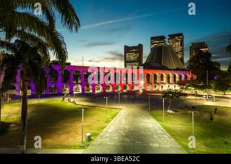 Lapa Aquädukt, beleuchtet mit violetten und rosa Farben am Abend mit hohen Bürogebäuden in der Innenstadt von Rio de Janeiro Stockfoto