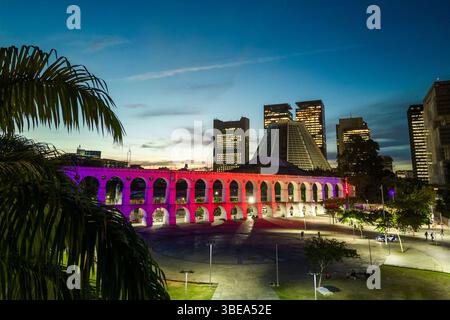 Lapa Aquädukt, beleuchtet mit violetten und rosa Farben am Abend mit hohen Bürogebäuden in der Innenstadt von Rio de Janeiro Stockfoto