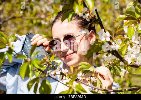 Eine Frau steht fröhlich zwischen blühenden Kirschbäumen, trägt Sonnenbrille und eine stilvolle Jacke, während sie sich im warmen Sonnenlicht eines Frühlings sonnt Stockfoto