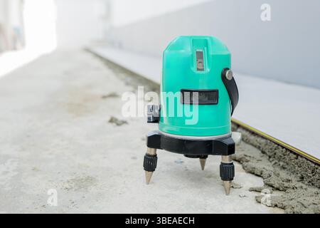 Eine grüne Laserwaage auf einem Stativ projiziert Strahlen über einen weißen und grauen Boden und sorgt so für Präzision Stockfoto