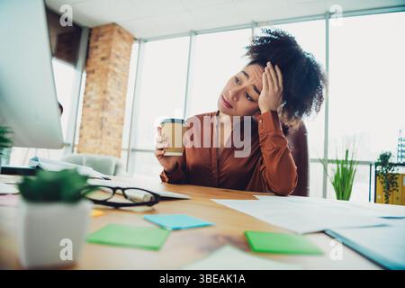 Junge professionelle Frau am modernen Schreibtisch, die Kaffee hält, während sie an einem stilvollen Arbeitsplatz mit industriellem Design arbeitet Stockfoto