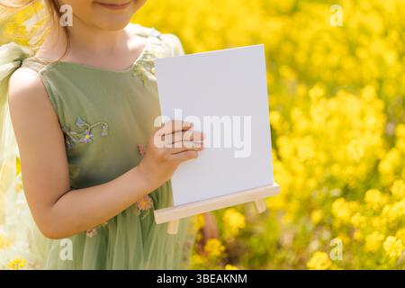 Beschnittene Aufnahme eines lächelnden Mädchens in grünem Kleid mit leerem Leintuch auf hölzernem Staffelei, umgeben von leuchtenden gelben Rapsblüten auf sonnigem Feld. Stockfoto