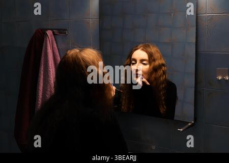 Eine junge Frau mit langen, welligen roten Haaren trägt Lippenstift in einem schwach beleuchteten Badezimmer auf, ihre Reflexion ist im Spiegel sichtbar. Stockfoto