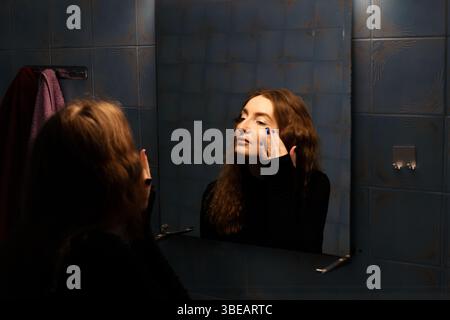 Eine junge Frau mit langen, welligen roten Haaren trägt Lippenstift in einem schwach beleuchteten Badezimmer auf, ihre Reflexion ist im Spiegel sichtbar. Stockfoto