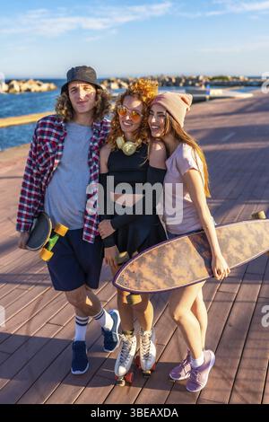 Drei glückliche Skater-Freunde posieren zusammen auf einer Promenade am Meer, halten Skateboards und Rollschuhe Stockfoto