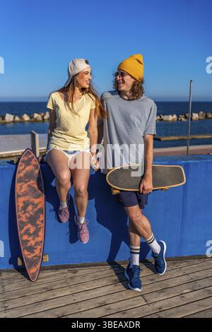 Glückliche junge Skater sitzen und stehen an einer blauen Wand am Meer, halten ihre Longboards und lächeln Stockfoto