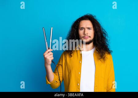 Junger Mann mit langen lockigen Haaren, der ein Stylingwerkzeug hält und Verwirrung vor einem leuchtend blauen Hintergrund ausdrückt Stockfoto