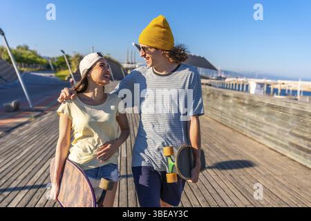 Glückliches junges Paar, das Hand in Hand an einem Pier entlang geht, Skateboards hält und an einem schönen Sommertag die Sonne genießt Stockfoto