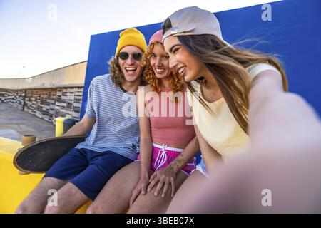 Drei fröhliche junge Freunde sitzen auf einem Dach und machen ein Selfie mit einem Skateboard in der Nähe Stockfoto