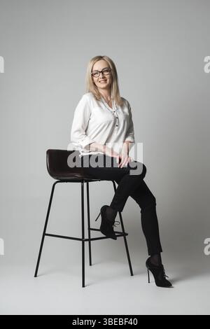 Lächelnde, schöne junge Frau mit Brille, die auf einem Stuhl sitzt, in einem Studio mit grauem Hintergrund. Vertriebsmitarbeiter, Manager, Agent, Assistent, Lehrer, Bolzen Stockfoto