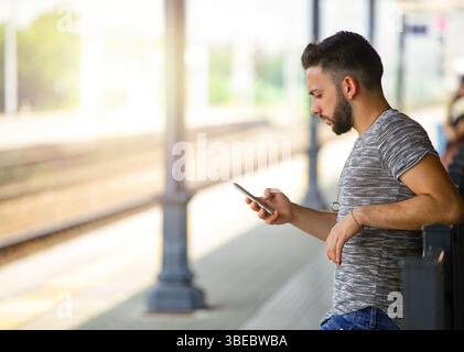 Lässig gekleideter junger Mann, der sich an einem Geländer lehnt und sein Smartphone benutzt, um in Verbindung zu bleiben, während er an einem hellen, sonnigen Tag auf einen Zug am Bahnhof wartet Stockfoto