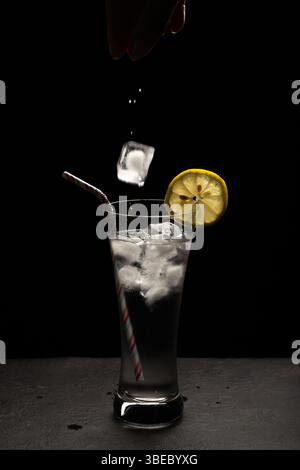 Ein Glas Wasser mit einer Zitronenscheibe darin. Ein Stroh ist im Glas. Eine Person gießt Eis in das Glas Stockfoto