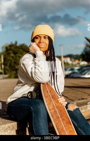 Eine junge lateinfrau mit Vitiligo in gelber Mütze und Jeans sitzt draußen, hält ein Skateboard und blickt nachdenklich nach oben, um die ess festzuhalten Stockfoto