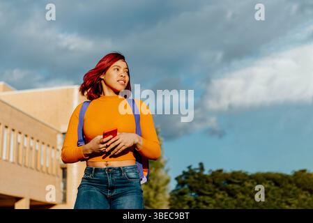 Eine junge Studentin mit rotem Haar und einem hellgelben Top geht über ihren College Campus, während sie ihr Smartphone ansieht Stockfoto