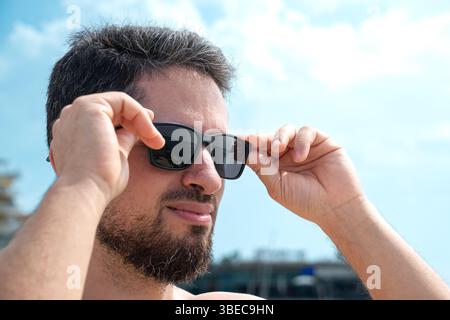 Junger erwachsener Mann, der seine Sonnenbrille auszieht und einen wunderschönen Strandblick und einen sonnigen blauen Himmel betrachtet Stockfoto